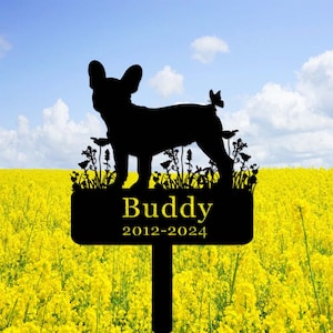 May include: Black metal silhouette of a French Bulldog with a butterfly above its tail, standing in a field of flowers. A black sign below reads "Buddy 2012-2024".