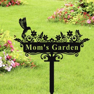 May include: Black metal garden sign with the text "Mom's Garden". The sign features a hummingbird, flowers, and a butterfly design. The sign is on a stake for easy placement in a garden or yard.