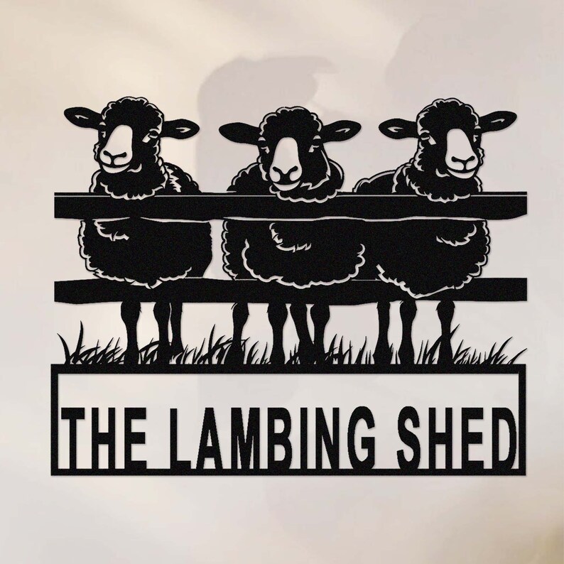 May include: Black metal sign featuring three sheep peering over a fence. The sign includes the text "THE LAMBING SHED" in a rectangular frame, with grass at the bottom. The sheep have white faces and black wool.