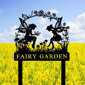 May include: Black metal silhouette of two fairies with wings, sitting on a flower bed with a butterfly, under a floral arch. The sign reads "FAIRY GARDEN".