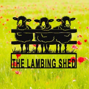 May include: Black metal sign featuring three sheep peering over a fence. The sheep have yellow faces and fluffy black wool. The sign reads "THE LAMBING SHED" in a rectangular frame, set against a blurred background of green grass and red flowers.