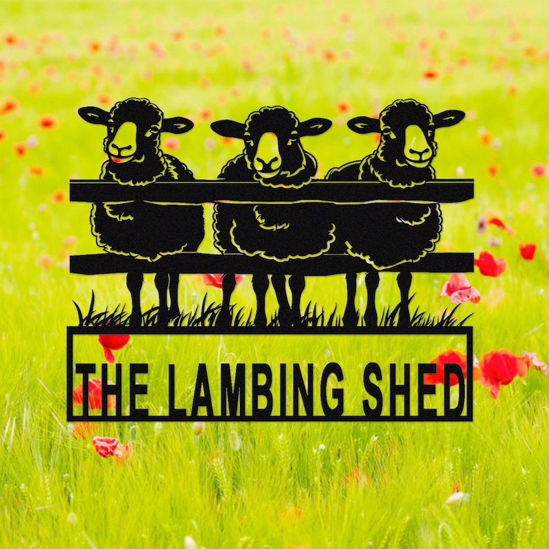 May include: Black metal sign featuring three sheep peering over a fence. The sheep have yellow faces and fluffy black wool. The sign reads "THE LAMBING SHED" in a rectangular frame, set against a blurred background of green grass and red flowers.