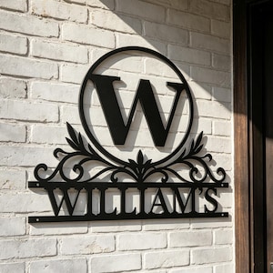 May include: A black metal wall sign featuring a large capital letter W centred within a circular frame. Below the circle, decorative scrollwork sits above a horizontal bar displaying the text WILLIAM'S in a bold, sans-serif font style.