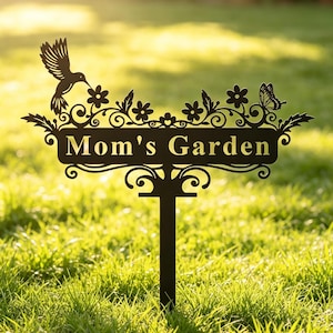Puede incluir: Un estaca de jardín de metal negro con una parte superior decorativa que presenta un colibrí, una mariposa y volutas florales. La placa rectangular central muestra el texto "Mom's Garden" en una fuente serif, sobre un poste vertical.