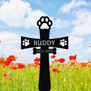 May include: Black metal cross with a paw print design at the top and bottom. The cross has the name "BUDDY" and the dates "2012-2024" engraved on it. The cross is in a field of red poppies.