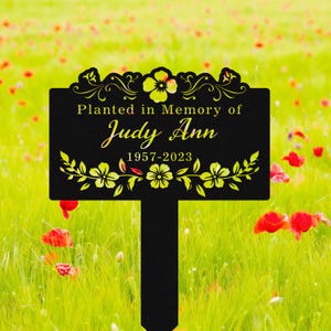 Peut inclure: Un piquet de jardin commémoratif en métal noir avec l'inscription "Planted in Memory of Judy Ann 1957-2023". Le piquet présente des motifs floraux jaunes et est planté dans un champ d'herbe verte et de fleurs rouges.