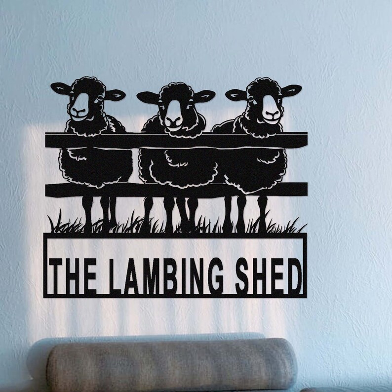 May include: Black metal wall art featuring three sheep peering over a fence. The sheep have fluffy wool and are set against a backdrop of grass. The words "THE LAMBING SHED" are displayed below the sheep.