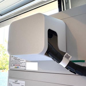 May include: A light grey electrical component with a curved top and a black cable emerging from it. A white label with a QR code is attached to the cable. The component is mounted on a larger, light grey unit.