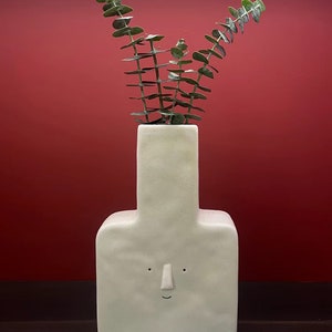 May include: White ceramic vase with a face and a square base, holding a sprig of green eucalyptus.