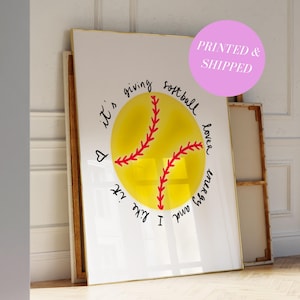 May include: A framed print featuring a yellow softball with red stitching. The text around the softball reads "It's giving softball lover energy and like it". The print has a gold frame and is leaning against a wall.