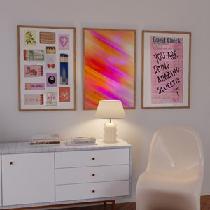 May include: Three framed prints on a white wall above a white dresser with three drawers. The left print features a collage of matchboxes with various designs. The middle print is a colourful abstract design with streaks of pink, orange, and yellow. The right print is a pink and white guest check with the text "You are doing amazing sweetie".