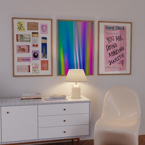 Può includere: Una cassettiera bianca con tre cassetti e pomelli dorati. La cassettiera ha un piano in piastrelle bianche con dei libri sopra. Ci sono tre quadri incorniciati sul muro sopra la cassettiera. L'immagine a sinistra è un collage di scatole di fiammiferi con vari disegni. L'immagine al centro è un'immagine astratta di linee verticali colorate. L'immagine a destra è un assegno di ospite rosa e bianco con il testo "You are doing amazing sweetie" scritto su di esso. Una lampada bianca con un paralume bianco si trova sulla cassettiera. Una sedia bianca si trova di fronte alla cassettiera.