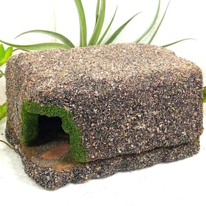 Pode incluir: Um abrigo retangular para animais com um exterior texturizado semelhante a cascalho em tons de castanho, cinzento e preto. A estrutura inclui uma única entrada em arco revestida com musgo artificial verde brilhante, criando uma aparência de caverna.
