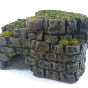 Op de afbeelding: Een miniatuur stenen muurstructuur met meerdere niveaus, voorzien van een donkerbruine, verweerde baksteentextuur en een boogvormige opening. De bovenzijden en kieren zijn bedekt met groen mos en kleine plukjes geelgroen gras.