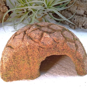 Könnte beinhalten: Ein kuppelförmiges Reptilienversteck mit einer strukturierten, rotbraunen Oberfläche, die an natürlichen Stein oder Erde erinnert. Die Struktur weist ein eingraviertes geometrisches Muster und eine bogenförmige Öffnung auf.