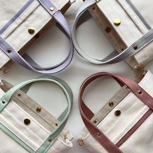 May include: Four canvas tote bags with pastel-coloured straps: lavender, grey, mint green, and dusty rose. Each bag has a cream-coloured body, gold hardware, and a rectangular shape. The straps are sewn onto the bag with matching coloured trim.