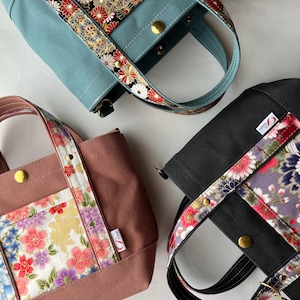 Op de afbeelding: Drie kleine draagtassen van blauwe, oudroze en zwarte canvas. Elke tas heeft een middenpaneel en handvatten versierd met een levendig bloemenpatroon in rood, wit, paars en goud. Elke tas is voorzien van een goudkleurige metalen drukknoopsluiting.