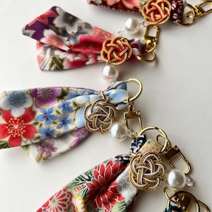May include: A collection of fabric keychains featuring colorful floral patterns in shades of red, blue, purple, and white. Each keychain includes a gold-toned metal lobster clasp, a decorative knotted cord charm, and a single white faux pearl bead.