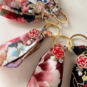 May include: Close-up of several fabric keychains with floral patterns in red, pink, and blue. Each keychain has a gold ring and a decorative knot with pearl and crystal accents. The keychains are on a white surface.