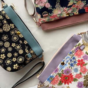 May include: Three rectangular crossbody bags with wrist straps and gold-tone hardware. One bag is black with gold floral patterns and a teal panel, one is navy with red and pink flowers, and one is white with colorful floral prints and a lavender panel.