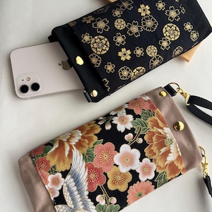 以下が含まれることがあります： 日本の花柄が施された2つの布製携帯電話ポーチ。1つは金色の花柄の黒色、もう1つは鶴と花柄です。どちらもリストストラップと金色のスナップ留めが付いています。1つのポーチには携帯電話が一部見えています。