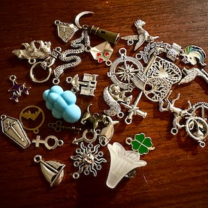 May include: A collection of silver charm pendants with various designs, including a house, a lighthouse, a compass, a tree, a cloud, a key, a sun, a clover, a unicorn, a deer, and a fish. The charms are arranged on a wooden surface.