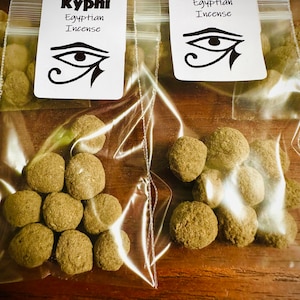 May include: Two clear plastic bags filled with small, round, brown incense cones. The bags are labeled "Kyphi Egyptian Incense" with an image of the Eye of Horus.