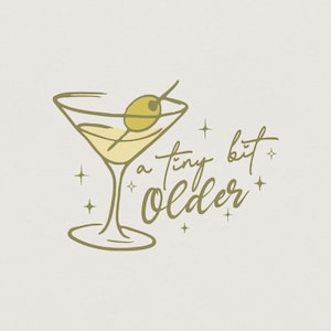 Puede incluir: Ilustración dorada de una copa de martini con una aceituna en un palillo. El texto "a tiny bit older" está escrito en una fuente de script debajo de la copa, con acentos de estrella. El diseño está sobre un fondo crema.