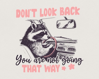 May include: A graphic illustration of a raccoon driving a car, with the text DON'T LOOK BACK in pink, and You are not going THAT WAY in black script, accompanied by two small pink flower icons at the bottom of the design.