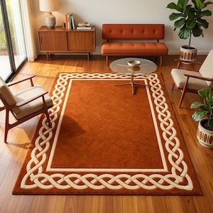 May include: A rectangular area rug featuring a solid burnt orange center field enclosed by a wide, cream-colored border decorated with an intricate, continuous Celtic knot pattern. The rug is positioned on a polished wooden floor surface.