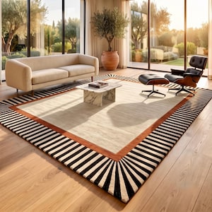 May include: A large rectangular area rug featuring a central cream-colored field, a solid burnt orange border, and an outer edge with a black and white striped sunburst pattern. The rug is positioned on a light wood floor in a bright living room.