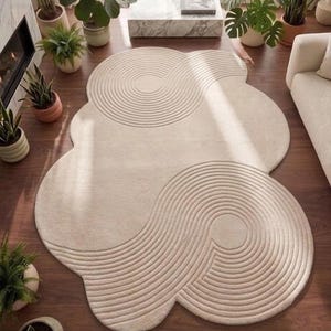 May include: An irregular, cloud-shaped area rug in a solid cream colour. The rug features a textured surface with carved, concentric circular patterns that mimic the appearance of raked sand in a zen garden, creating a flowing, organic design.