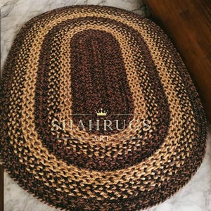 以下が含まれることがあります： 茶色とベージュの色合いの楕円形の編み込みラグ。ラグは、より明るい茶色とベージュの編み込みボーダーに囲まれた、中央の濃い茶色の部分が特徴です。「SHAH RUGS ART」というテキストがラグに表示されています。