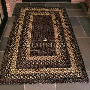 May include: A rectangular braided rug in shades of brown and tan. The rug features a central rectangular design surrounded by a border of alternating colors. The text "SHAHRUGS ART" is visible on the rug.