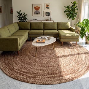 May include: A contemporary living room with an olive green corner sofa, a circular woven jute rug, and an oval coffee table. The room is decorated with framed artwork and potted plants, creating a stylish and inviting space.