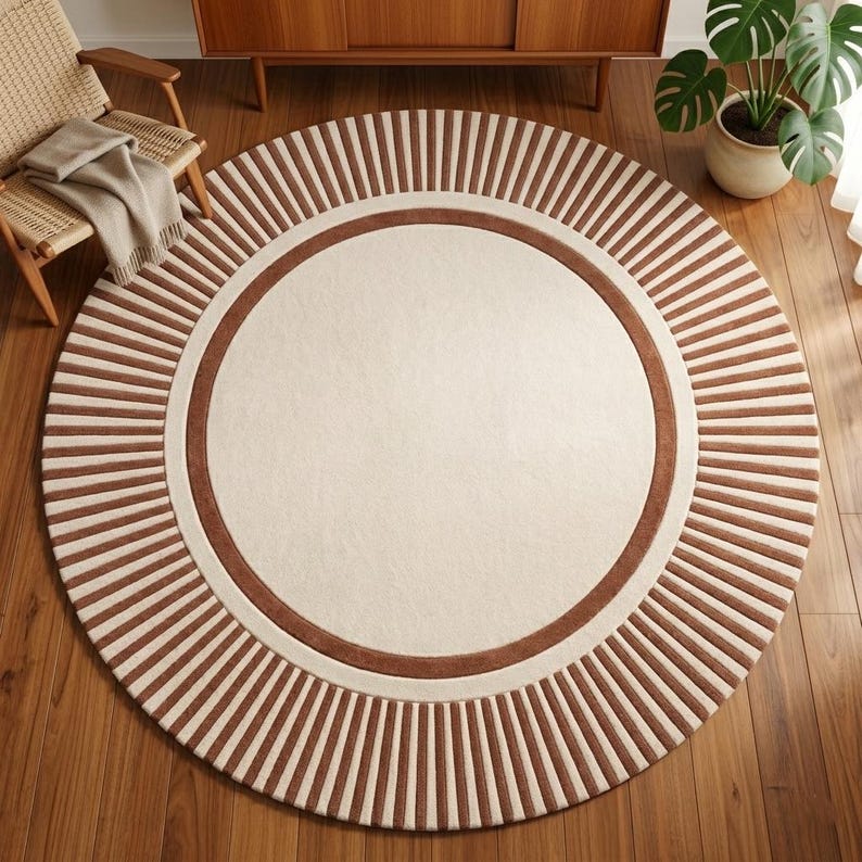 Puede incluir: Una alfombra redonda con un centro crema y un borde marr&oacute;n. El borde exterior presenta rayas verticales marrones que irradian desde el centro. La alfombra est&aacute; sobre un suelo de madera, con una silla y una planta en el fondo.