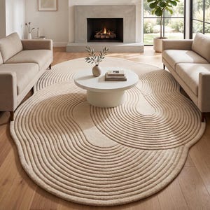 Op de afbeelding: Een beige, cirkelvormig vloerkleed met een uniek, vloeiend ontwerp. Het kleed heeft concentrische, gebogen lijnen die een golfachtig patroon creëren. Het staat in een woonkamer met een witte salontafel en twee beige banken.