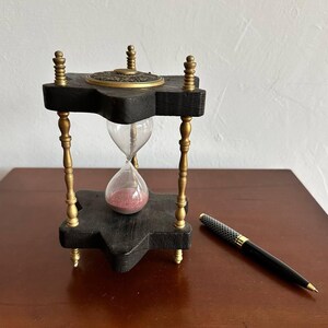 Puede incluir: Un reloj de arena de madera con una parte superior y una base de latón. El reloj de arena está lleno de arena roja y se encuentra sobre una mesa de madera. Un bolígrafo negro está situado junto al reloj de arena.