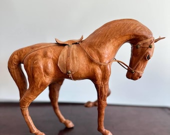 Puede incluir: Una figurita de caballo de cuero marrón con una silla de montar y una brida. El caballo está de pie sobre una superficie marrón oscura.