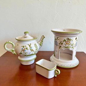 May include: A collection of ceramic kitchenware, including a teapot, a small rectangular container with a handle, and a decorative candle holder. The items are white with green trim and floral and bird designs. The teapot has a lid and a curved spout.