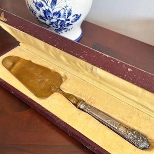 以下が含まれることがあります： 金色の刃と装飾的な銀色のハンドルが付いたヴィンテージケーキサーバー。バーガンディ色のベルベットの裏地が付いた箱に入っています。刃にはエッチングされた花の模様があり、ハンドルには詳細な模様があります。