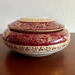 May include: A round porcelain trinket box with a lid. The box is decorated with a burgundy base and gold floral patterns. The lid has a white center with gold floral patterns.
