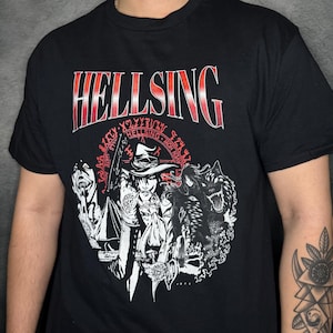 Puede incluir: Camiseta negra con un gráfico rojo de "Hellsing". El gráfico presenta un personaje con un sombrero y un lobo, rodeado de un círculo de texto.