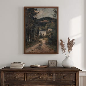 May include: A framed landscape painting depicts a dirt road leading to a white building nestled among trees. The painting is in a dark wooden frame. A dark wooden dresser holds books, candles, and a vase with dried plants.
