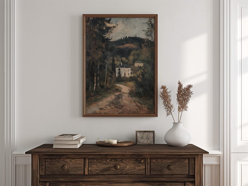 May include: A framed landscape painting depicts a dirt road leading to a white building nestled among trees. The painting is in a dark wooden frame. A dark wooden dresser holds books, candles, and a vase with dried plants.