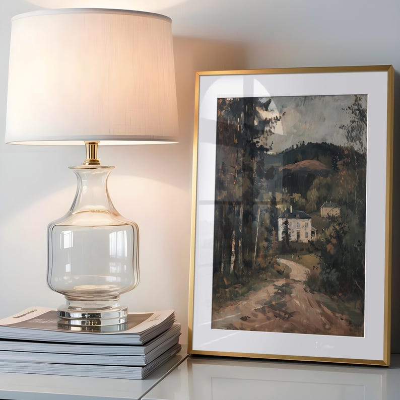 May include: A clear glass table lamp with a white lampshade and gold accents sits on a stack of magazines. Beside it is a framed landscape painting with a gold frame, depicting a house and trees.