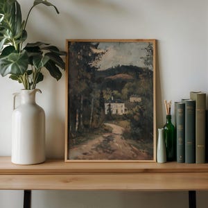 May include: A framed landscape painting depicts a winding road leading to a white house nestled among trees. The artwork is in a light wooden frame, displayed on a wooden surface with a white vase, green bottles, and books.