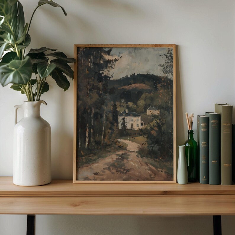 May include: A framed landscape painting depicts a winding road leading to a white house nestled among trees. The artwork is in a light wooden frame, displayed on a wooden surface with a white vase, green bottles, and books.