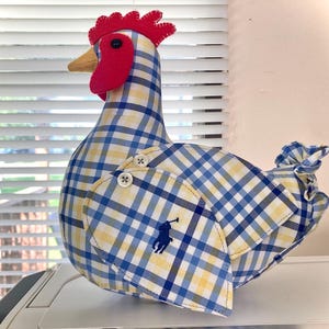 May include: A decorative chicken figurine made from fabric with a blue, yellow, and white plaid pattern. It features a red felt comb and wattle, a yellow beak, and a blue logo. The chicken has button details and a ruffled tail.