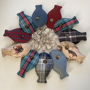 May include: A collection of ten fabric fish-shaped keychains arranged in a circle. Each keychain features a central button and is attached to a metal split ring. The fabrics include various plaid patterns, solid blue and red, and two with fishing fly prints.